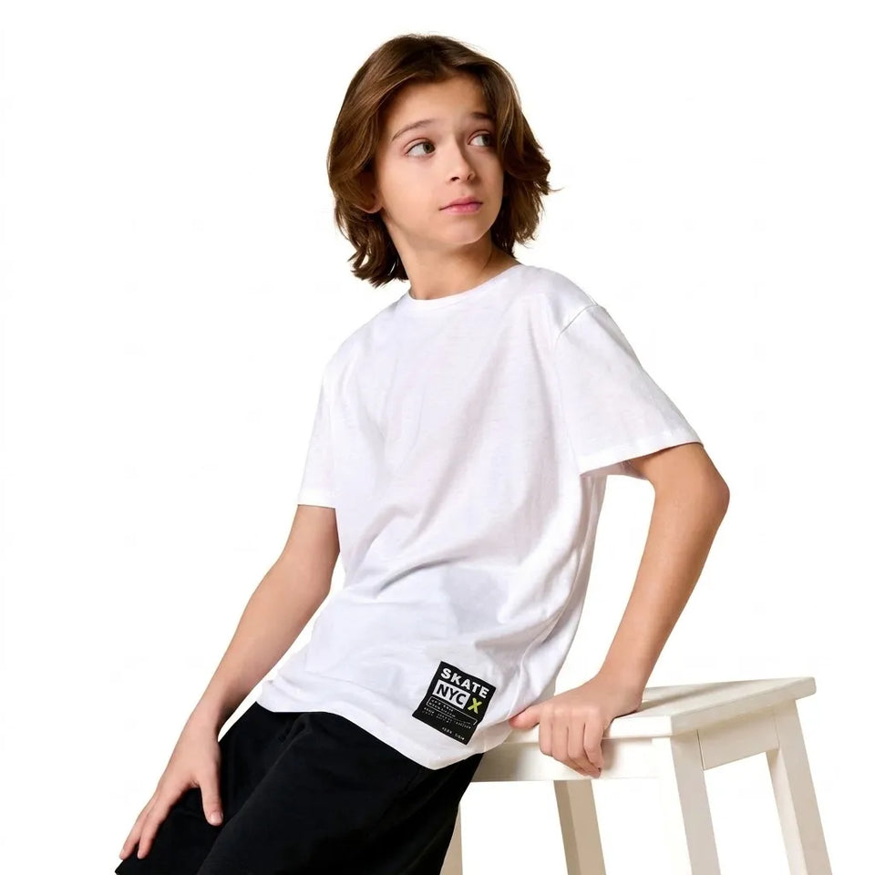A child model wearing a white short-sleeve crewneck t-shirt with a black 'BRITISH CIGAR' patch on the lower left hem, leaning on a white wooden stool against a white background.