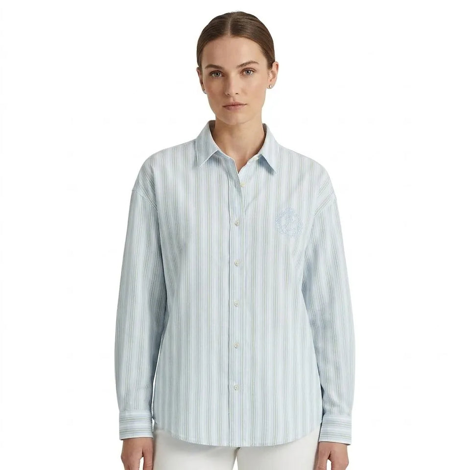 A woman modeling a light blue and white vertically striped long-sleeve button-up shirt with a subtle embroidered crest on the left chest, set against a plain white background.