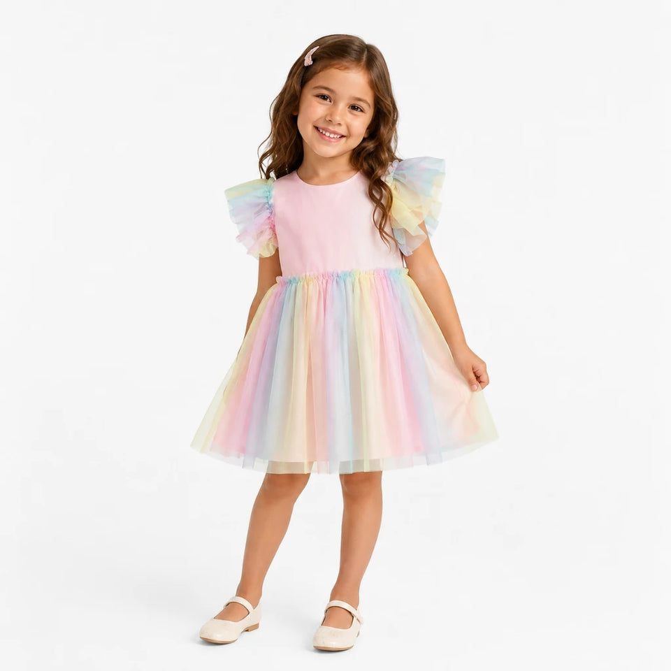 A young girl modeling a pastel tulle dress with a light pink bodice and a layered rainbow tulle skirt, paired with cream Mary Jane shoes, against a plain white studio background.