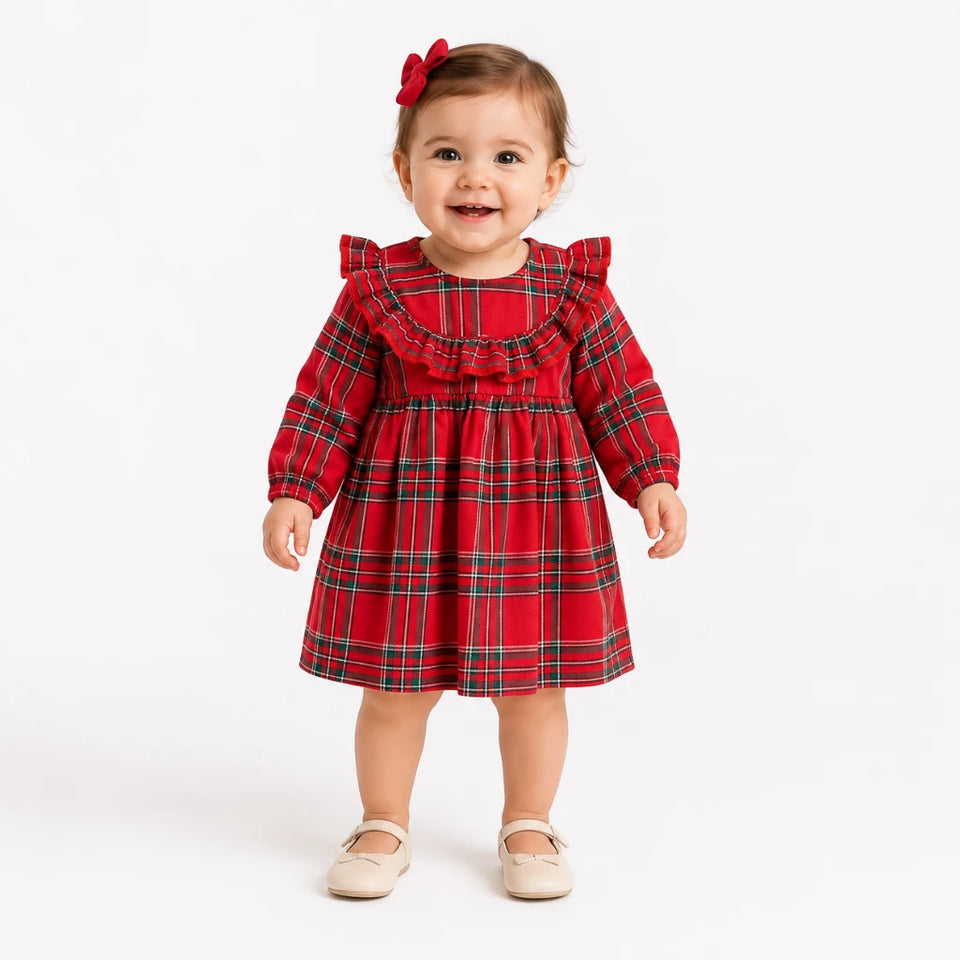 A toddler girl wearing a red tartan plaid dress with ruffled shoulders and a matching red bow in her hair, standing against a plain white studio background.