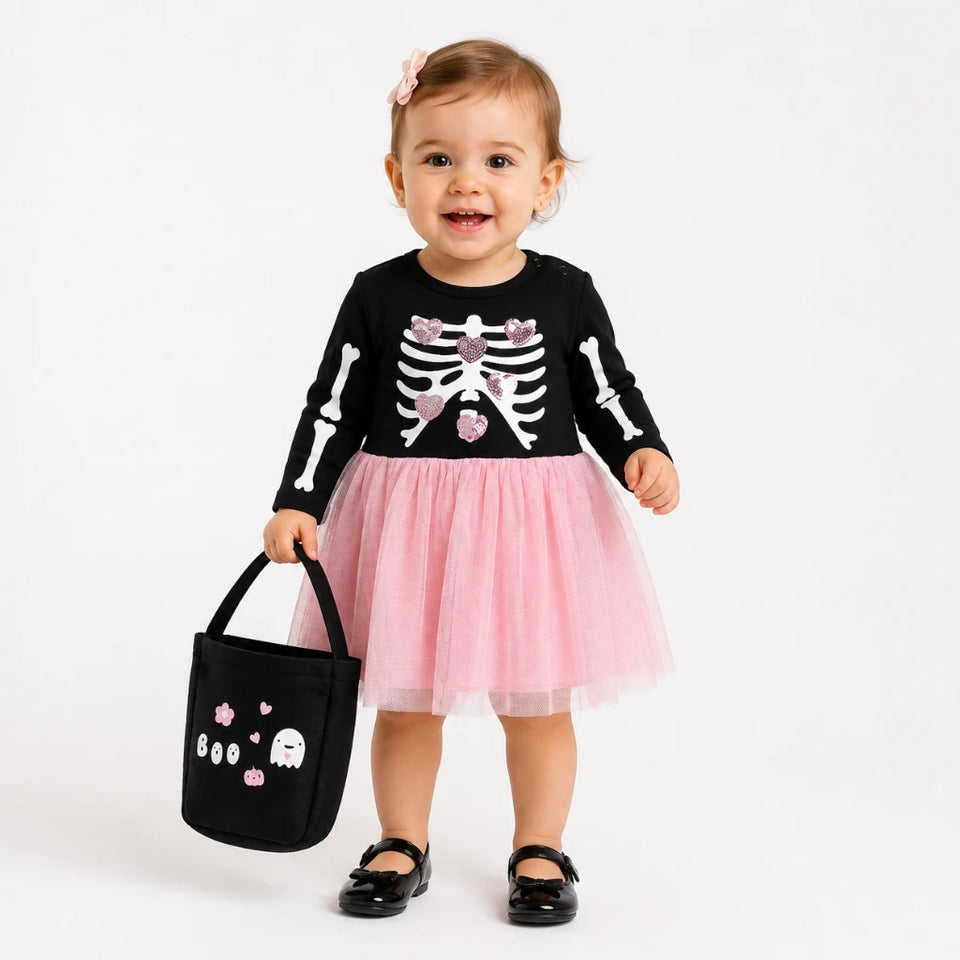 A toddler stands against a plain light background wearing a black long-sleeve top with a white skeleton print and a pink tulle skirt, holding a black tote bag with ghost and heart graphics.