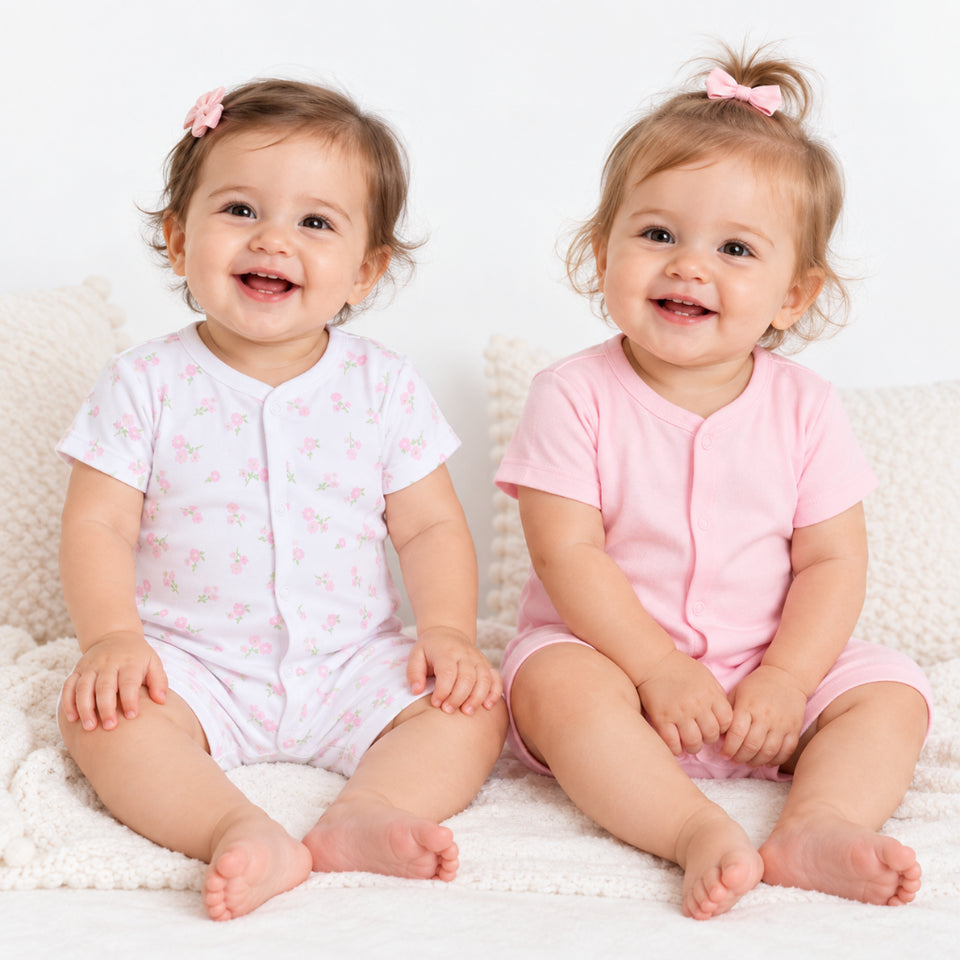 Two smiling babies sitting side by side on a white textured blanket. The baby on the left wears a white short-sleeve bodysuit with a small pink bow pattern, and the baby on the right wears a solid light pink short-sleeve bodysuit.