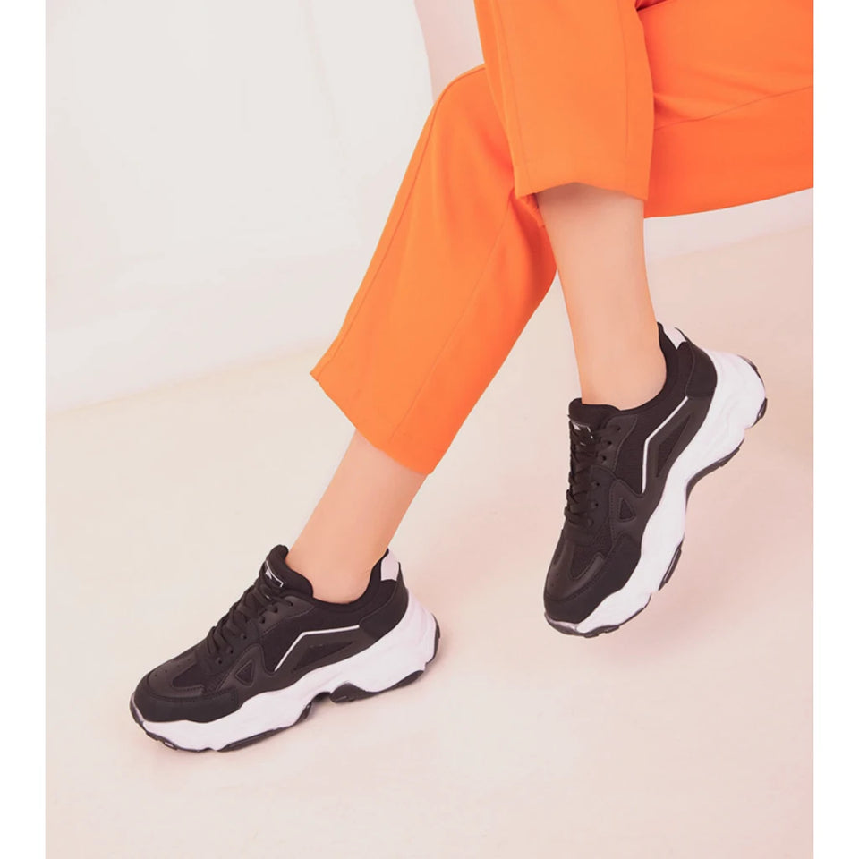 A person wearing black chunky platform sneakers with white midsoles and white side stripes, paired with bright orange pants, shown against a clean light beige studio background.