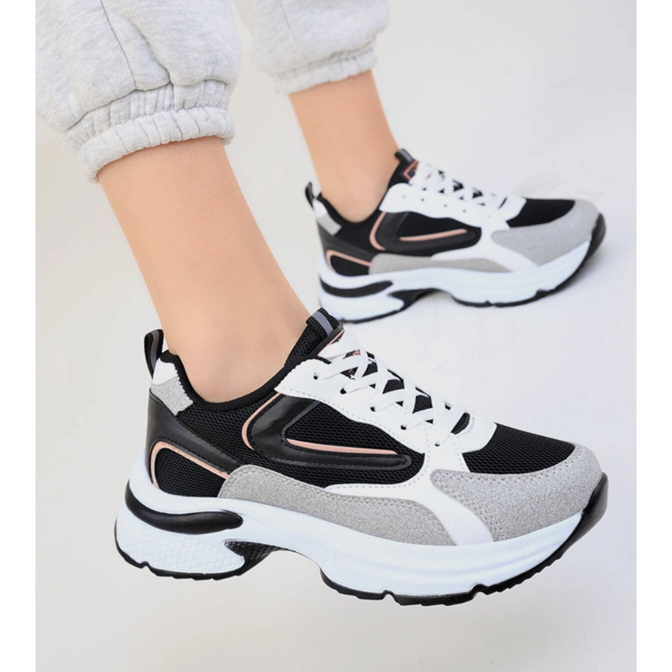 A pair of chunky low-top sneakers worn on a person's feet, shown against a plain light background. The shoes are primarily black and white with light gray panels and a subtle pink accent line.