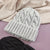 A light gray cable-knit beanie hat with a ribbed cuff, styled on a lavender surface with a beige blanket in the background and a dark gray beanie partially visible at the bottom.