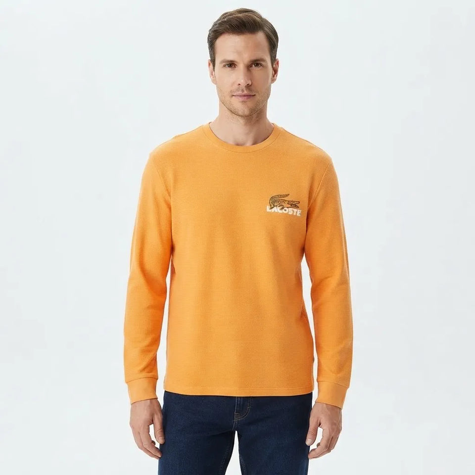 A man wearing a bright orange long-sleeve crewneck sweatshirt with a small embroidered Lacoste crocodile logo on the left chest, paired with dark blue jeans, against a plain light gray background.
