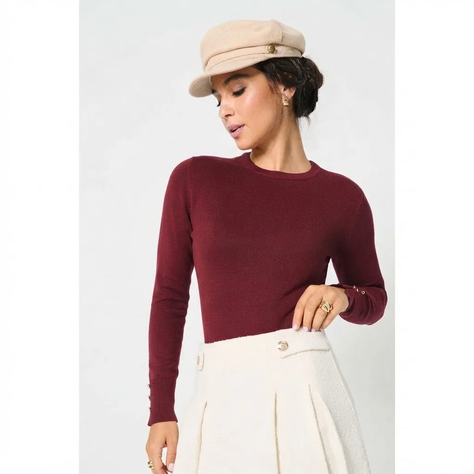 A woman wearing a fitted long-sleeve crewneck top in deep burgundy, paired with a cream pleated skirt and a beige newsboy cap, photographed against a light gray studio background.