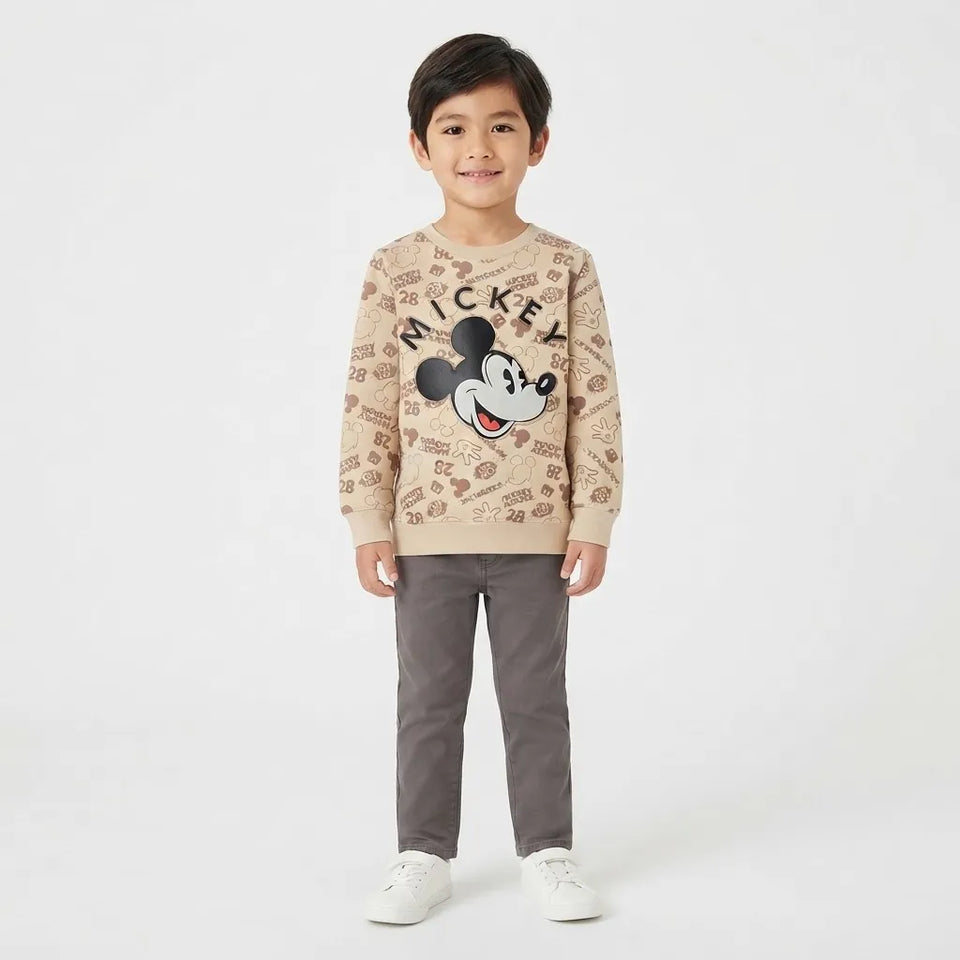 A young boy stands against a plain white studio background wearing a beige long-sleeve crewneck sweatshirt with a large Mickey Mouse graphic and 'MICKEY' text on the front, paired with dark gray pants and white sneakers.
