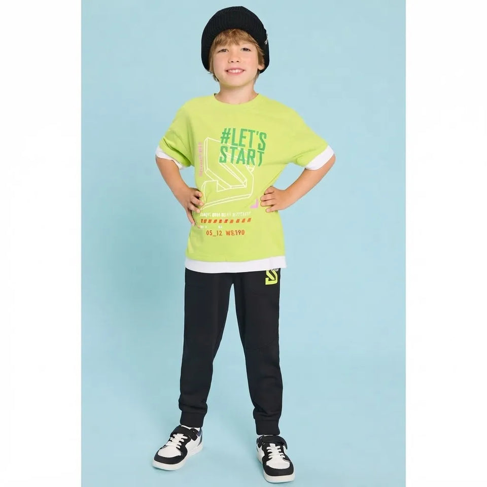 A young boy modeling a bright lime green graphic t-shirt with white and green details, paired with black sweatpants and a black beanie, standing against a light blue background.