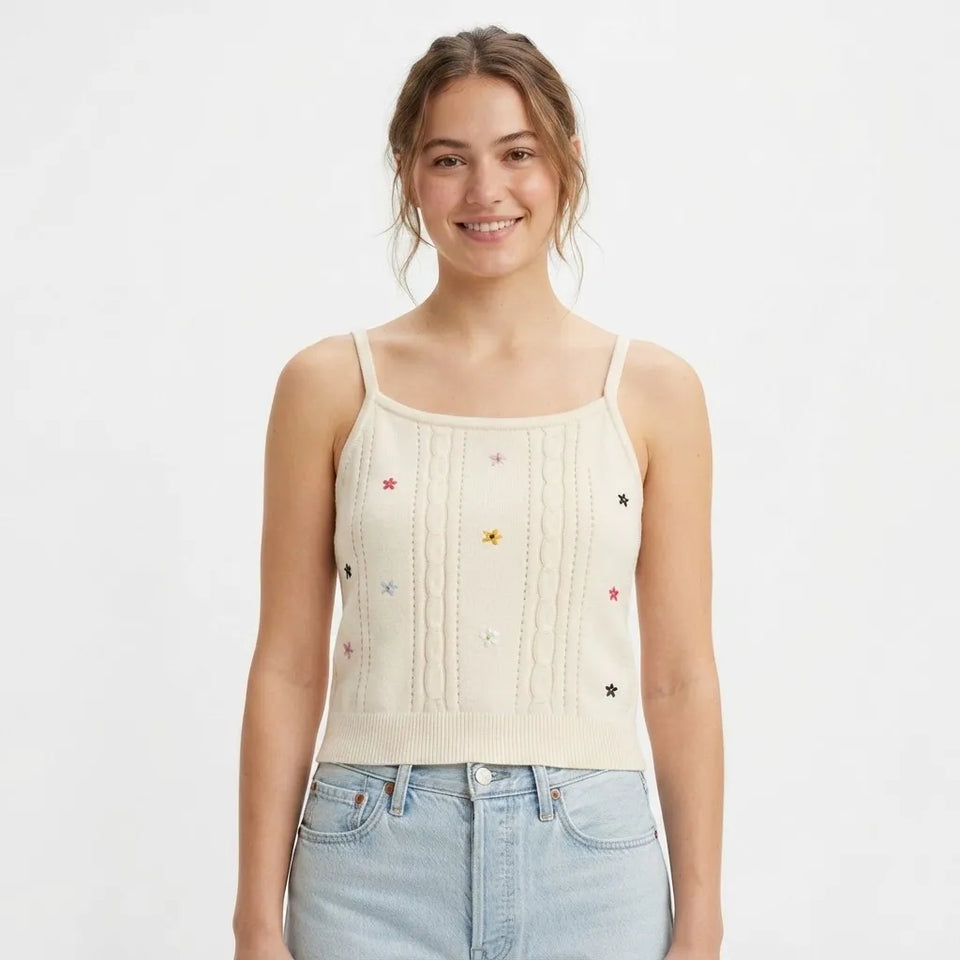 A woman wearing a cream-colored knit camisole top with small embroidered stars in multiple colors, paired with light-wash blue jeans, against a plain white studio background.