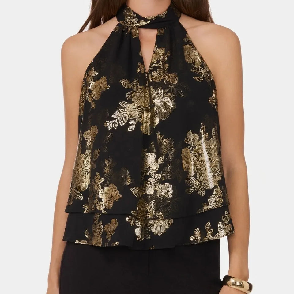 A woman wearing a black halter-neck blouse with a metallic gold floral pattern, featuring a keyhole cutout at the collar and a layered ruffle hem, set against a plain light gray background.