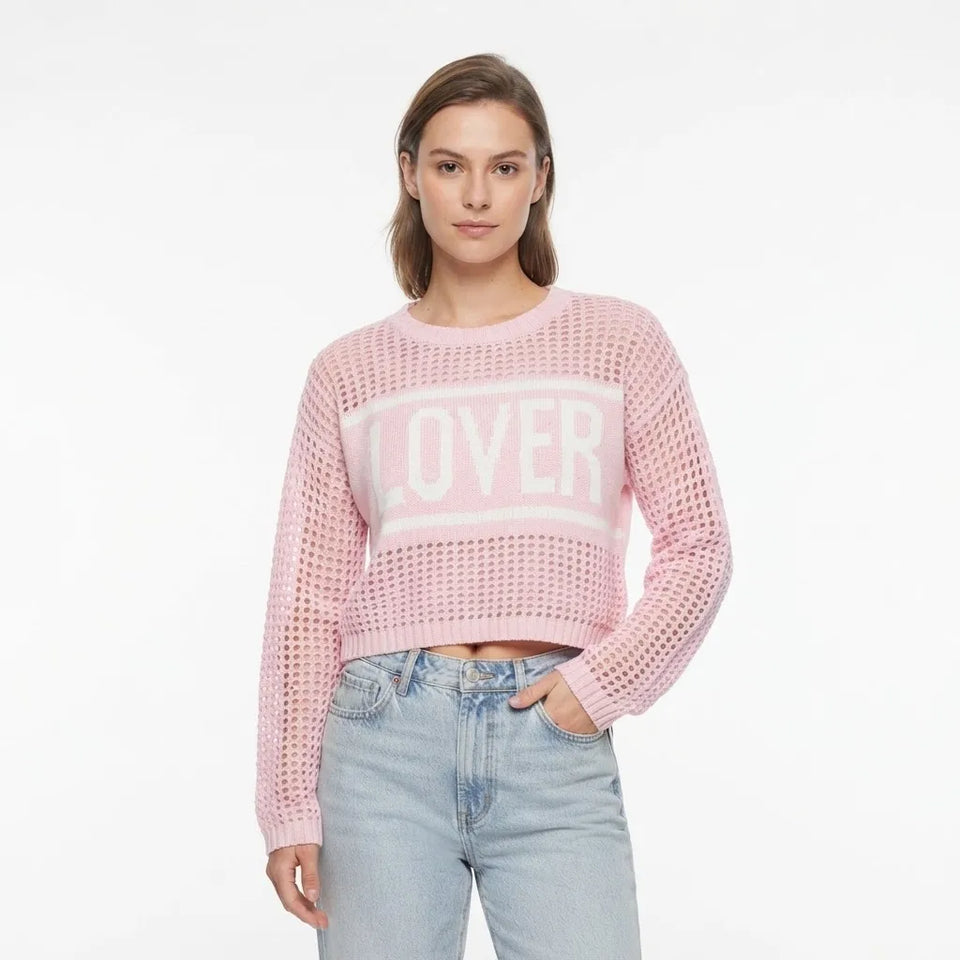 A woman modeling a light pink cropped knit sweater with a white 'LOVER' graphic across the chest, paired with light blue jeans, against a plain white background.