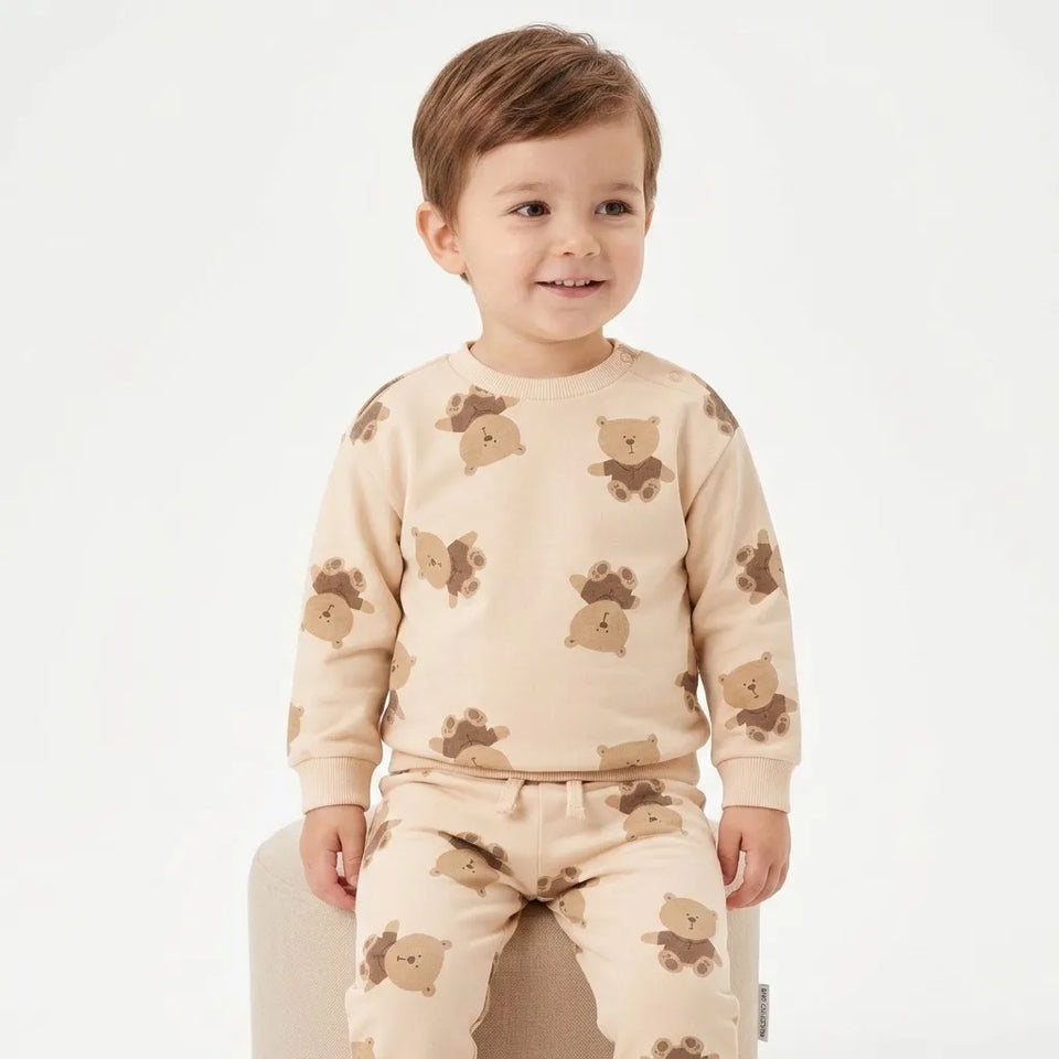 A young child wearing a beige long-sleeve pajama set with a brown teddy bear pattern, sitting on a light-colored chair against a plain white background.