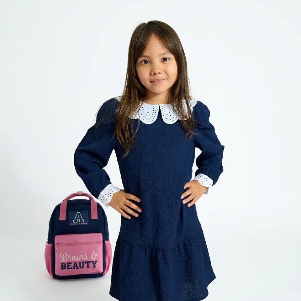 A young girl with long brown hair stands wearing a navy blue long-sleeve dress with a white lace Peter Pan collar and white lace cuffs. She is posing next to a pink and navy blue backpack with the words 'Brains & BEAUTY' on the front pocket, set against a plain white studio background.