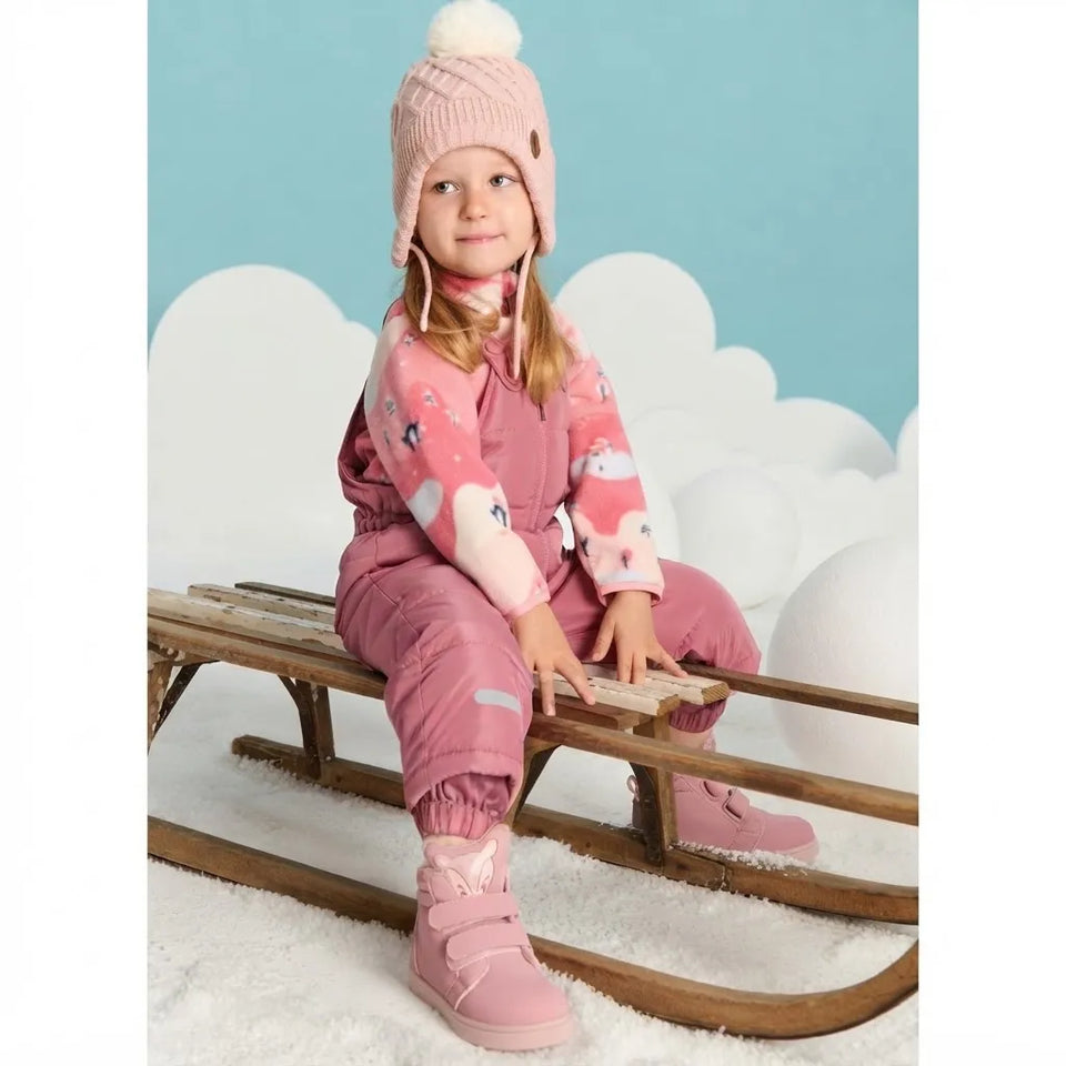 A young girl wearing a pink winter ski suit, matching hat, and boots sits on a wooden sled against a light blue background with white cloud shapes.