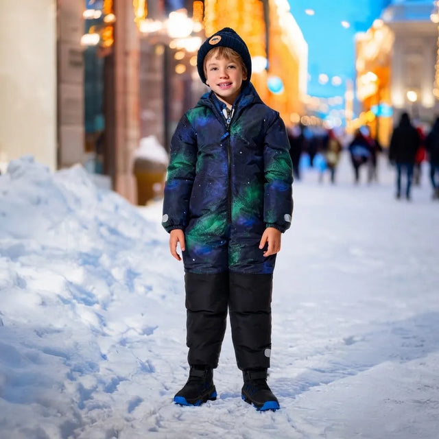 A young boy stands in the snow wearing a dark winter parka with a green and blue nebula pattern, black pants, and a black beanie, set against a blurred city street at night with warm lights.