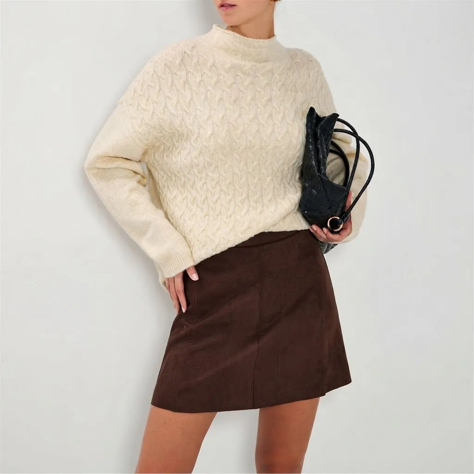 A model wearing a cream cable-knit mock-neck sweater and a brown mini skirt, holding a black textured shoulder bag against a plain light gray background.