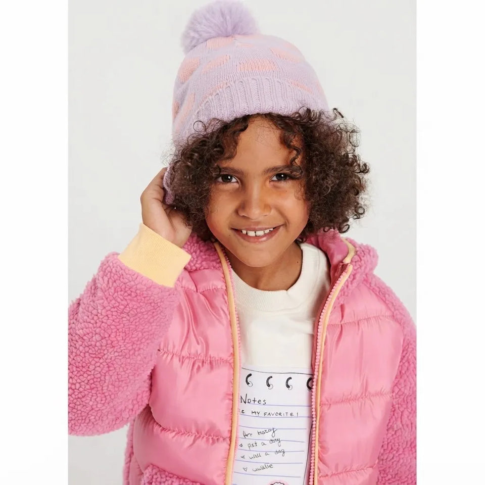 A young child wearing a pink puffer jacket with a fuzzy pink sleeve, a white t-shirt, and a light purple knit beanie with a pom-pom.