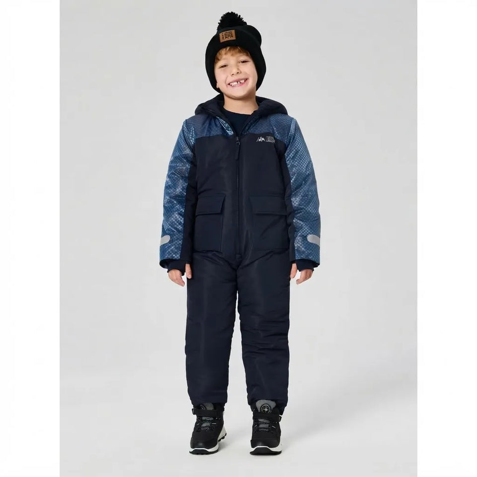 A young boy wearing a dark navy and blue patterned snowsuit with a black beanie and black sneakers, standing against a light gray studio background.