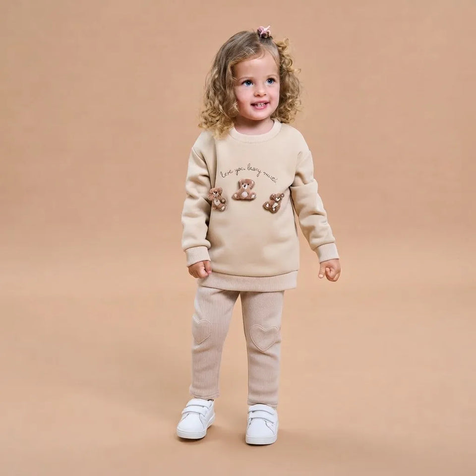 A toddler girl stands against a solid beige studio background wearing a matching beige sweatshirt and jogger pants set. The sweatshirt features a graphic of three brown teddy bears and the text 'Love you, baby love.