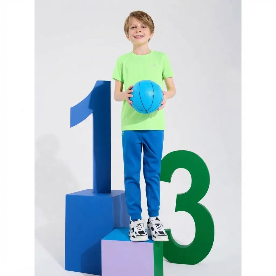 A young boy stands on a small purple block, smiling and holding a blue basketball. He is wearing a bright lime green t-shirt, blue sweatpants, and white sneakers.