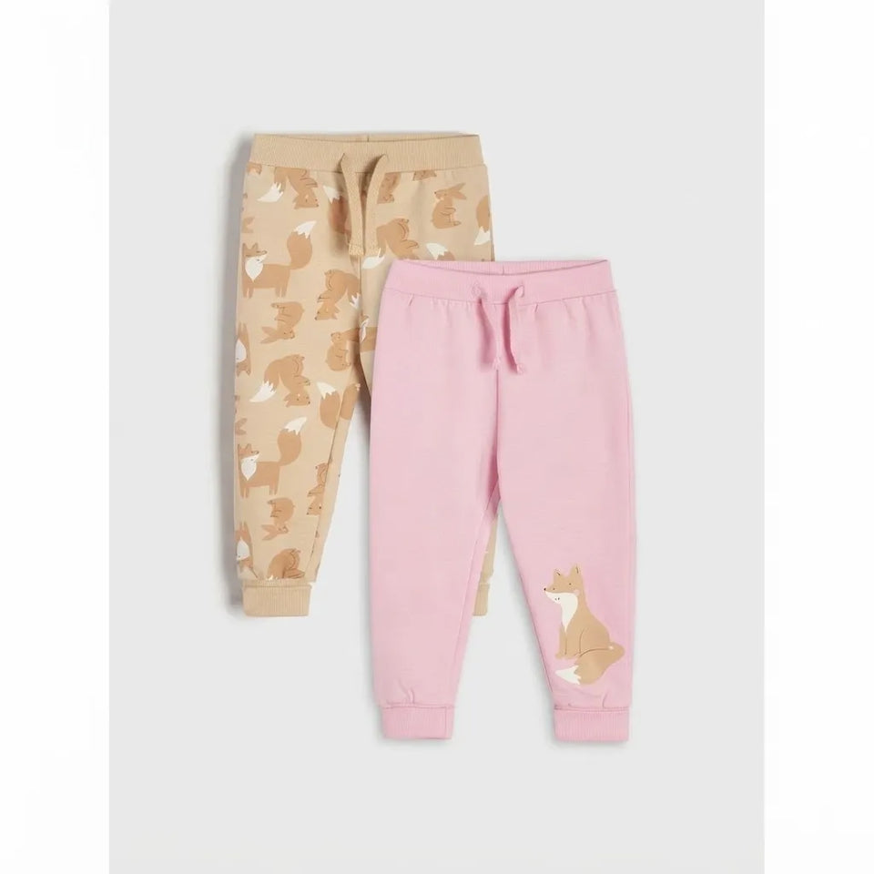 Two pairs of children's jogger sweatpants laid flat on a light gray background. The pair in back is beige with an allover fox print, and the pair in front is light pink with a large fox graphic on the lower left leg.