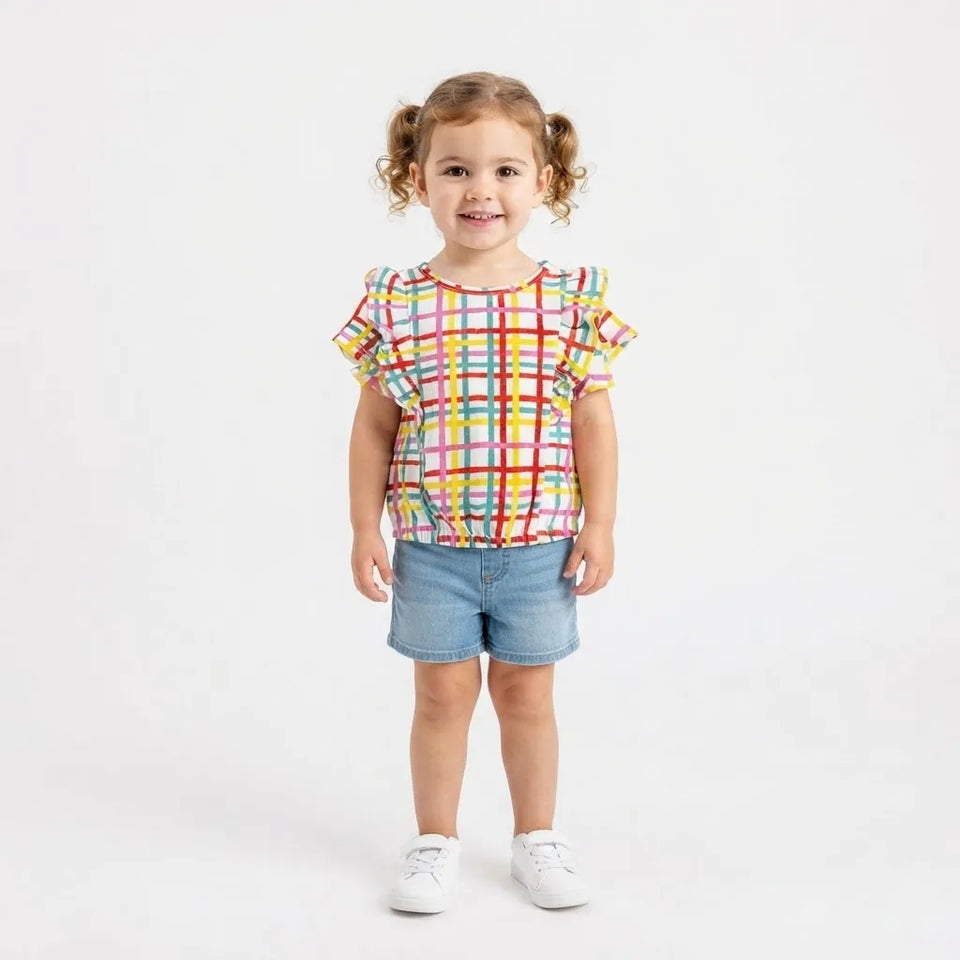 A young girl with curly brown hair in pigtails stands against a white studio background wearing a colorful plaid short-sleeve top, denim shorts, and white sneakers.