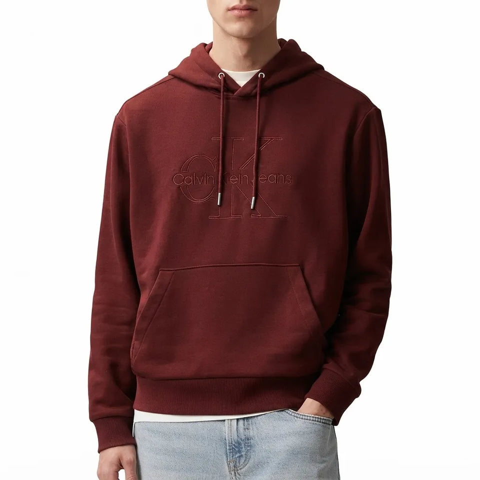 A man wearing a dark red/burgundy Calvin Klein hoodie with a tonal embroidered logo on the chest, paired with light blue jeans, against a plain white background.
