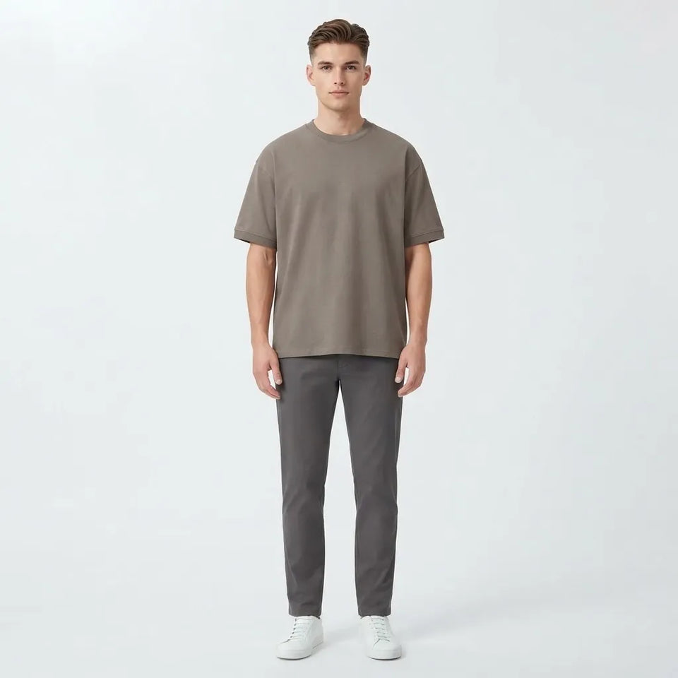 A man modeling a taupe short-sleeve crewneck t-shirt and dark gray trousers against a light gray studio background.