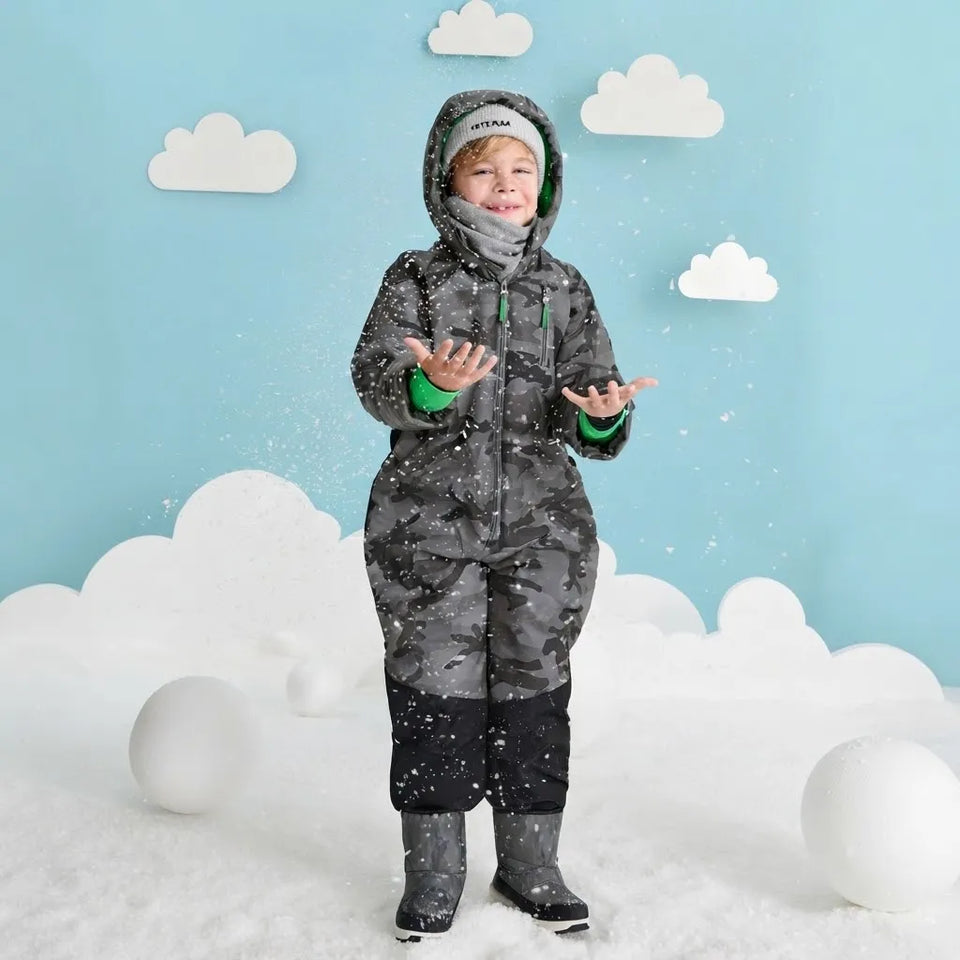 A child wearing a full-body gray camouflage snowsuit with green accents, a hood, and black boots stands in a snowy studio setup with white cloud props and a light blue background.