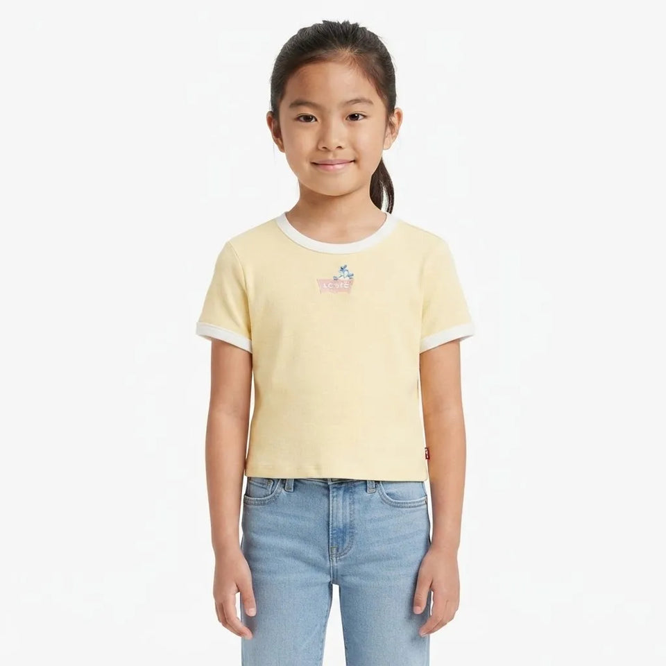 A young girl with her hair in a ponytail stands against a plain white background wearing a pale yellow ringer t-shirt with white ribbed trim and a small pink graphic on the chest, paired with light blue jeans.