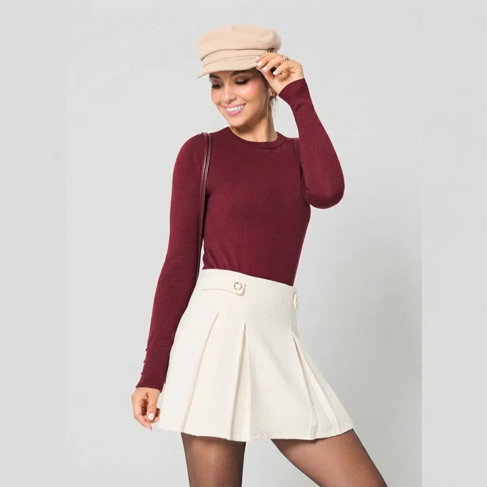 A woman posing in a studio setting wearing a burgundy long-sleeve knit top, an off-white pleated mini skirt, a beige newsboy cap, and black tights.