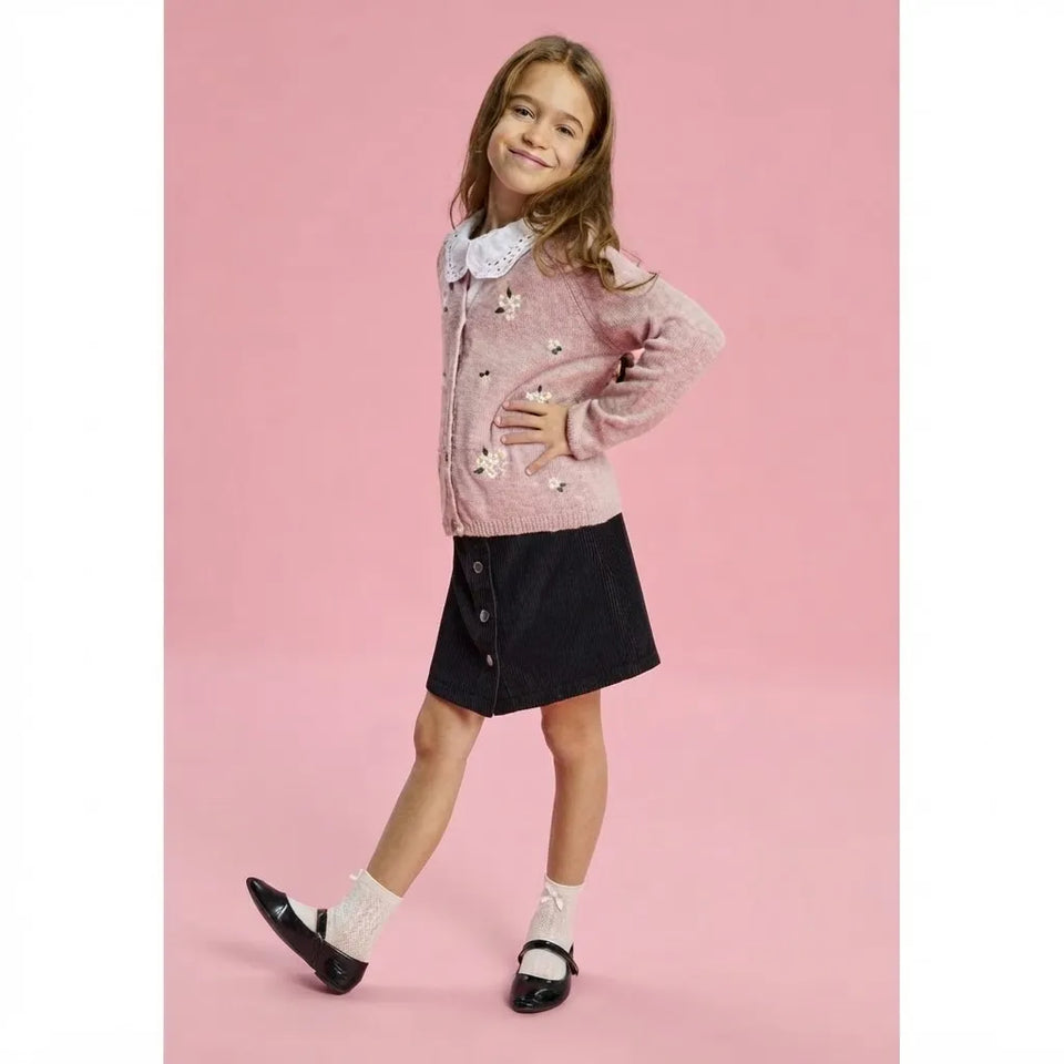 A young girl modeling a pink knit cardigan with white floral embroidery and a white lace collar, paired with a black skirt, white socks, and black Mary Jane shoes, set against a solid pink studio background.