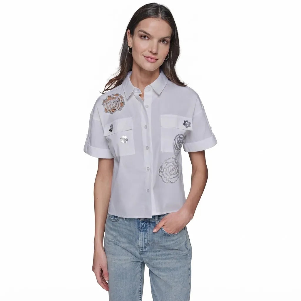 A woman modeling a white short-sleeve button-up shirt with embroidered floral details and silver sequins on the chest, paired with light-wash blue jeans against a plain white background.