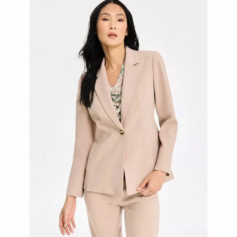 A woman modeling a tailored beige blazer with matching trousers, worn over a patterned blouse, against a plain light background.