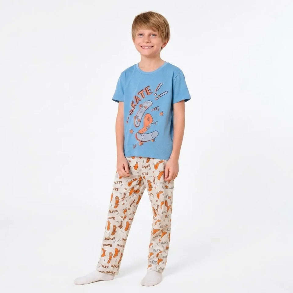 A young boy modeling a two-piece pajama set consisting of a light blue t-shirt with a skate graphic and cream-colored pants with an orange and black dog print.