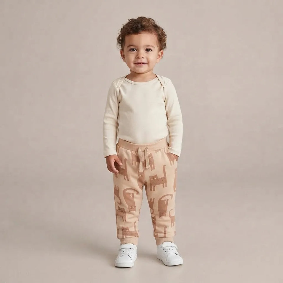 A toddler standing against a plain beige studio background, wearing a cream long-sleeve top and beige patterned jogger pants with white sneakers.