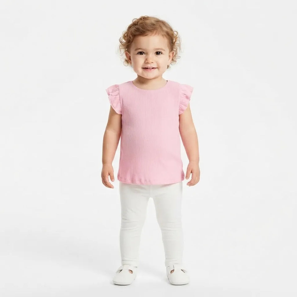A toddler girl with curly light brown hair stands against a plain white studio background wearing a light pink ribbed short-sleeve top with ruffled edges, white pants, and white shoes.