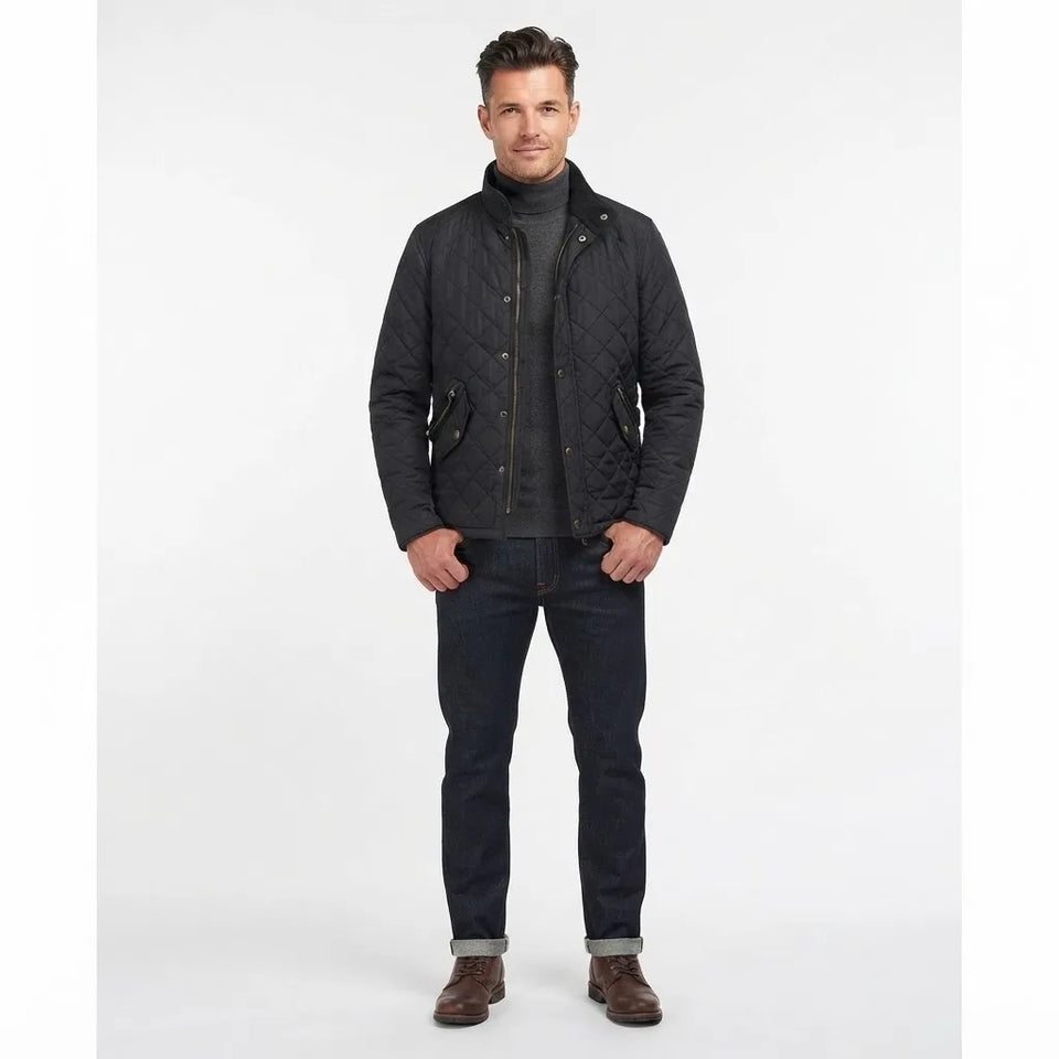A man modeling a black quilted jacket over a dark gray turtleneck sweater, dark blue jeans, and brown leather shoes, standing against a plain white studio background.