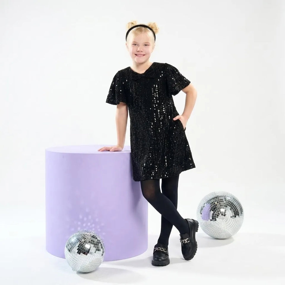 A young girl with blonde hair in a bear-ear headband stands next to a lavender cylinder, wearing a black sequined short-sleeve dress, black tights, and black shoes. Two silver disco balls are on the floor against a white studio background.