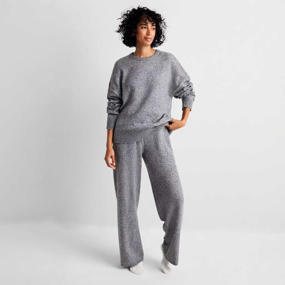 A woman modeling a matching gray knit lounge set consisting of a crewneck sweatshirt and wide-leg pants, photographed against a light gray studio background.