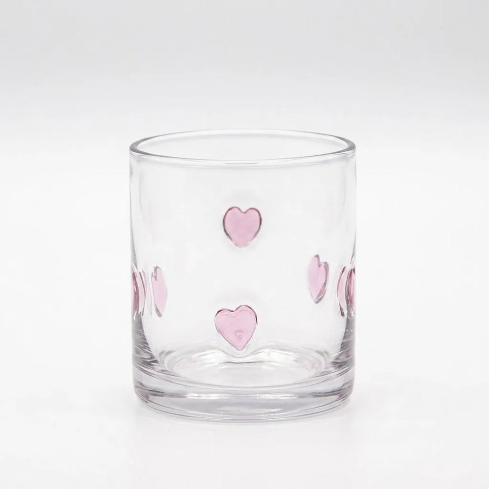 A clear glass tumbler with a circular base decorated with small pink heart shapes outlined in metallic silver, set against a seamless white background.