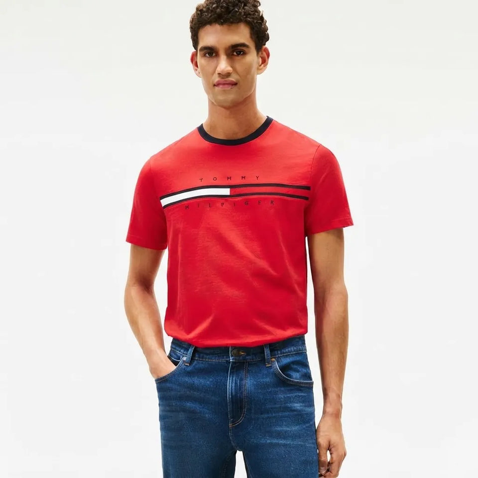 A man wearing a red Tommy Hilfiger short-sleeve t-shirt with a black, white, and red chest logo stripe, paired with blue jeans, against a plain white background.