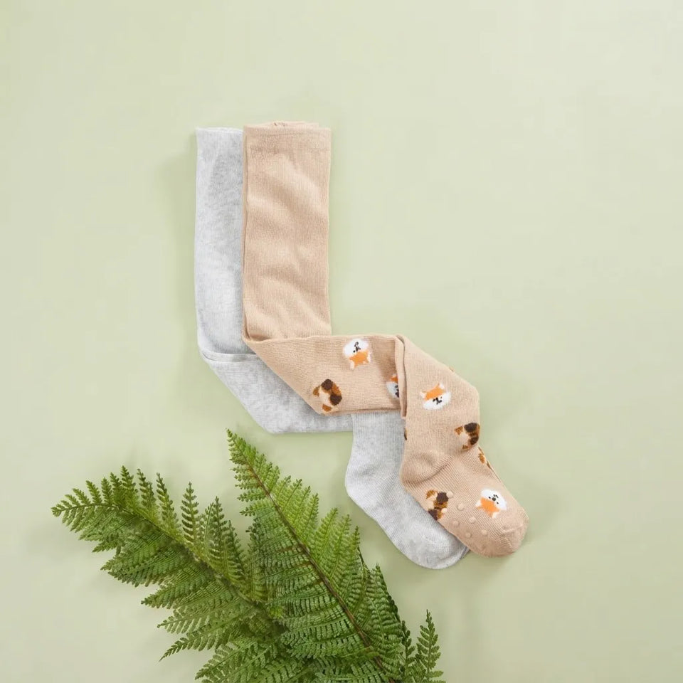 Three pairs of socks arranged on a pale green background with a green fern leaf at the bottom left. The socks include two solid light gray pairs and one tan pair with a small fox print.