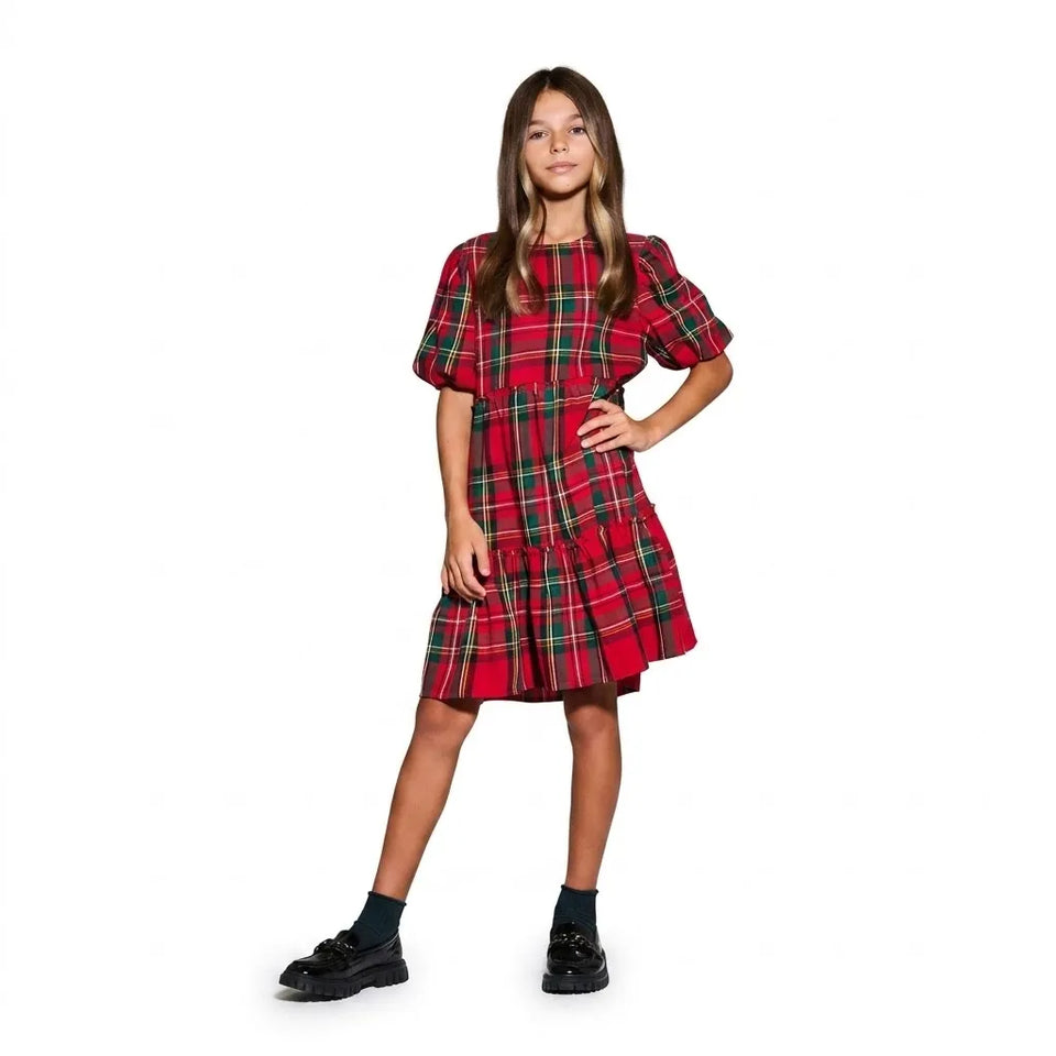 A young girl modeling a red tartan plaid dress with green and yellow stripes, featuring short puff sleeves and a tiered skirt, paired with black socks and shoes, against a plain white background.
