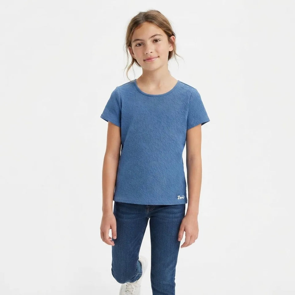 A young girl modeling a short-sleeve crewneck t-shirt in a heathered medium blue color, paired with dark blue jeans, against a plain white studio background.