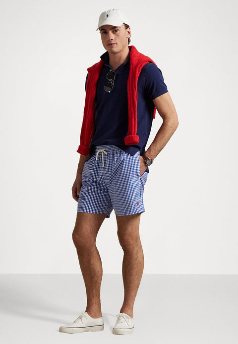 Man wearing a navy polo shirt, blue checkered shorts, red vest, and white cap on a white background