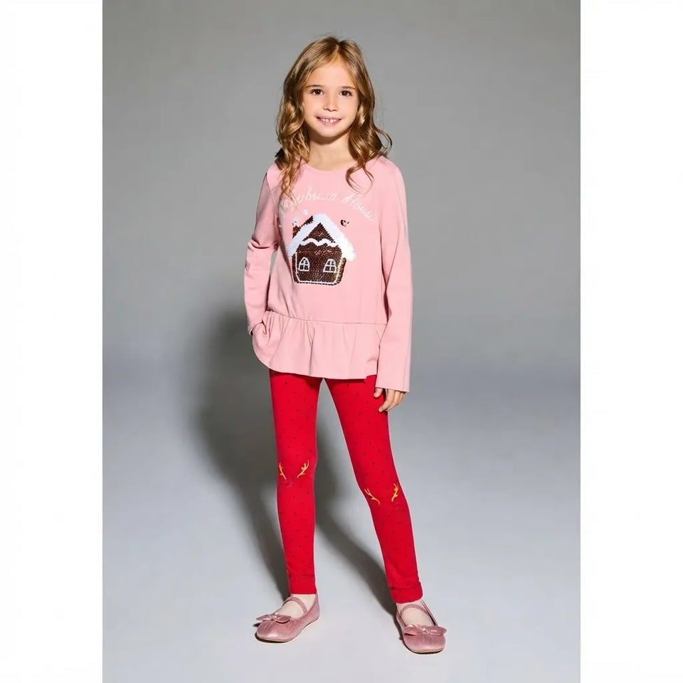 A young girl stands against a gray studio background wearing a light pink long-sleeve top with a gingerbread house graphic and red leggings with small gold details.