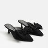 A pair of black satin pointed-toe mule heels with large bow details on the top, displayed on a clean white background.