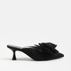 A single black pointed-toe mule heel with a large layered fabric flower detail across the vamp, shown from a side profile against a clean white background.